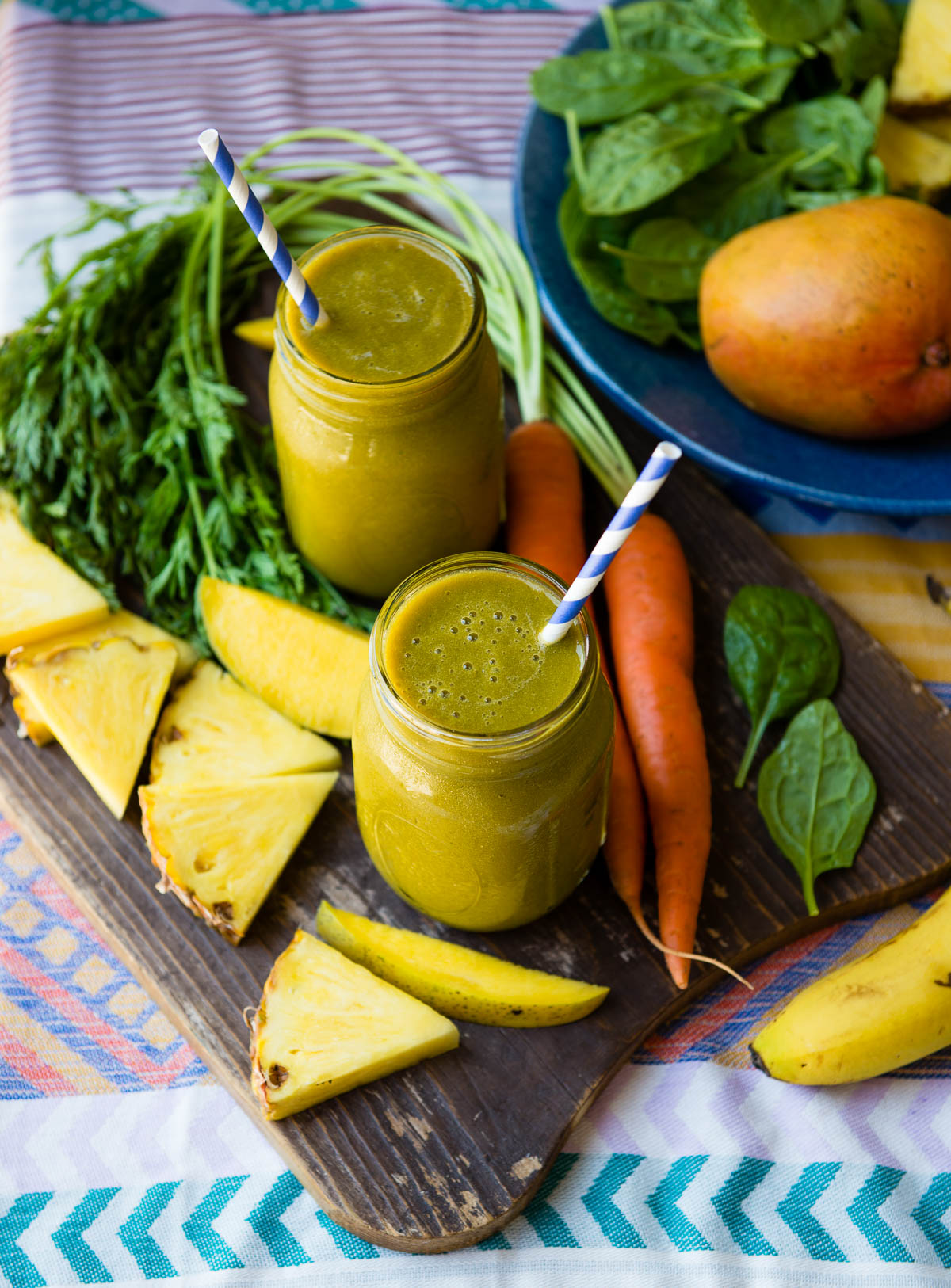 2 glass jars of carrot tops smoothie with papers straws, surrounded by fresh carrots, mango and pineapple slices.