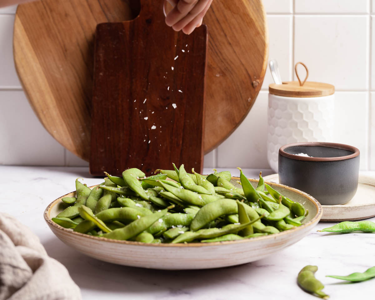 plate of cooked bean pods and a white hand sprinkling coarse salt over top.